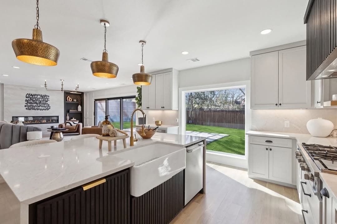 The kitchen at 13739 Hughes Lane, a stone island with a farmhouse sink under three hammered brass pendants, open through to the living room at the far end.
