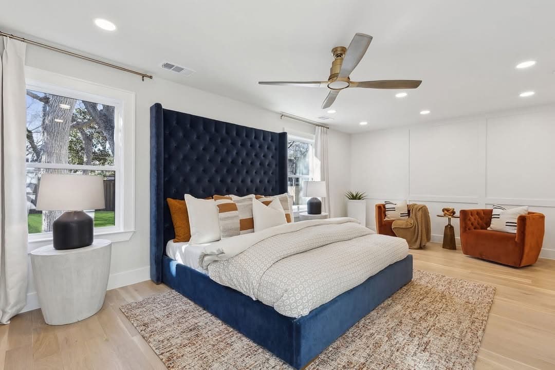 The primary bedroom at 13739 Hughes Lane, a navy buttoned headboard the height of the wall with two rust leather chairs in the bay beyond it.