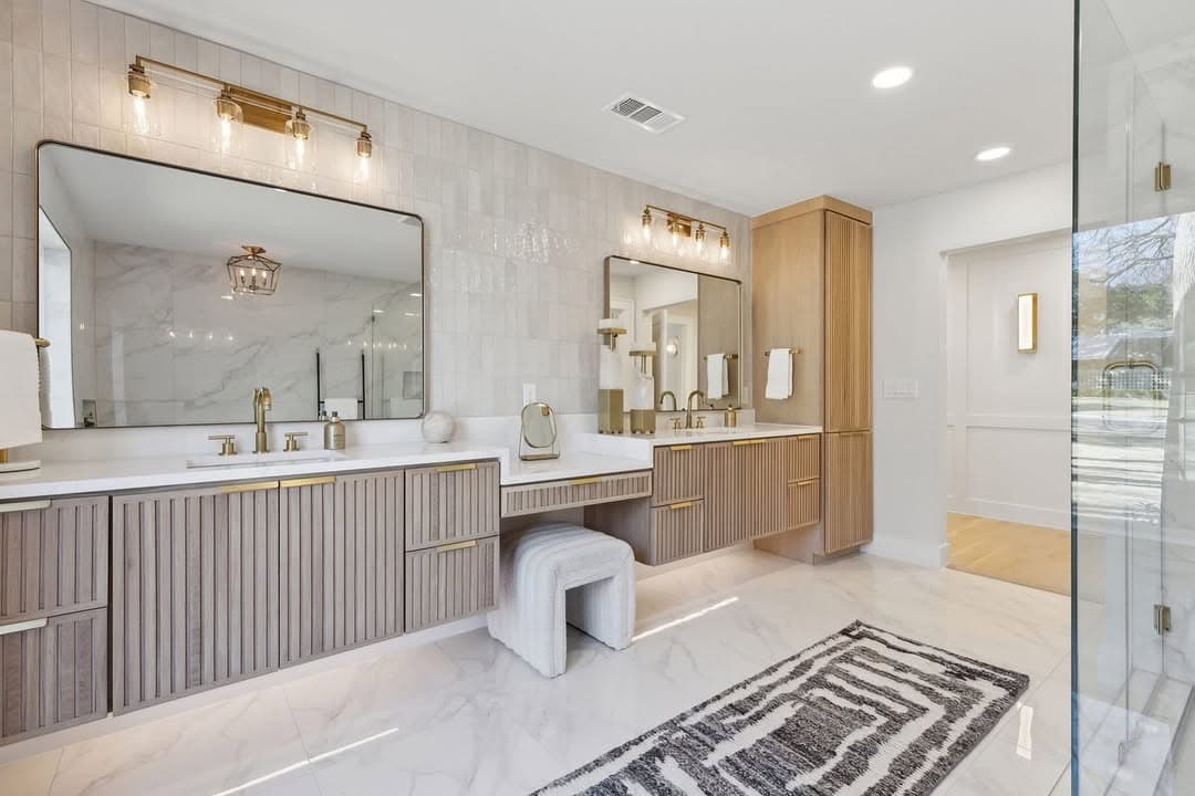 The primary vanity at 13739 Hughes Lane, twin fluted vanities under brass sconces with a fitted stool between them on marble tile.