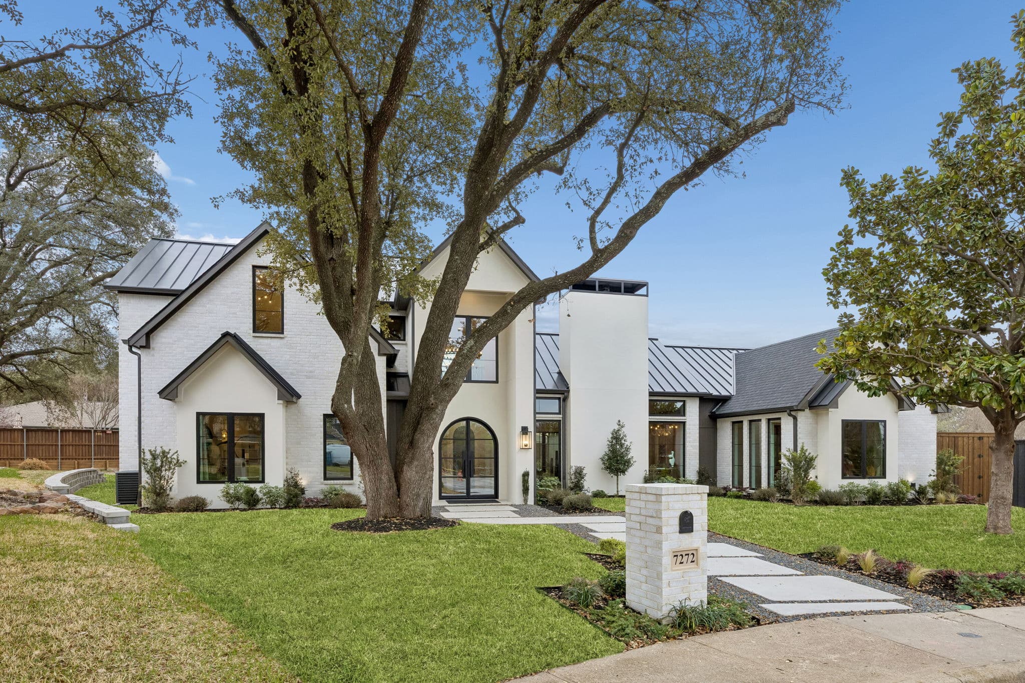 The front of 7272 Tangleglen, a white rendered house under a dark standing seam roof, a broad oak shading the lawn and a stone path to the door.
