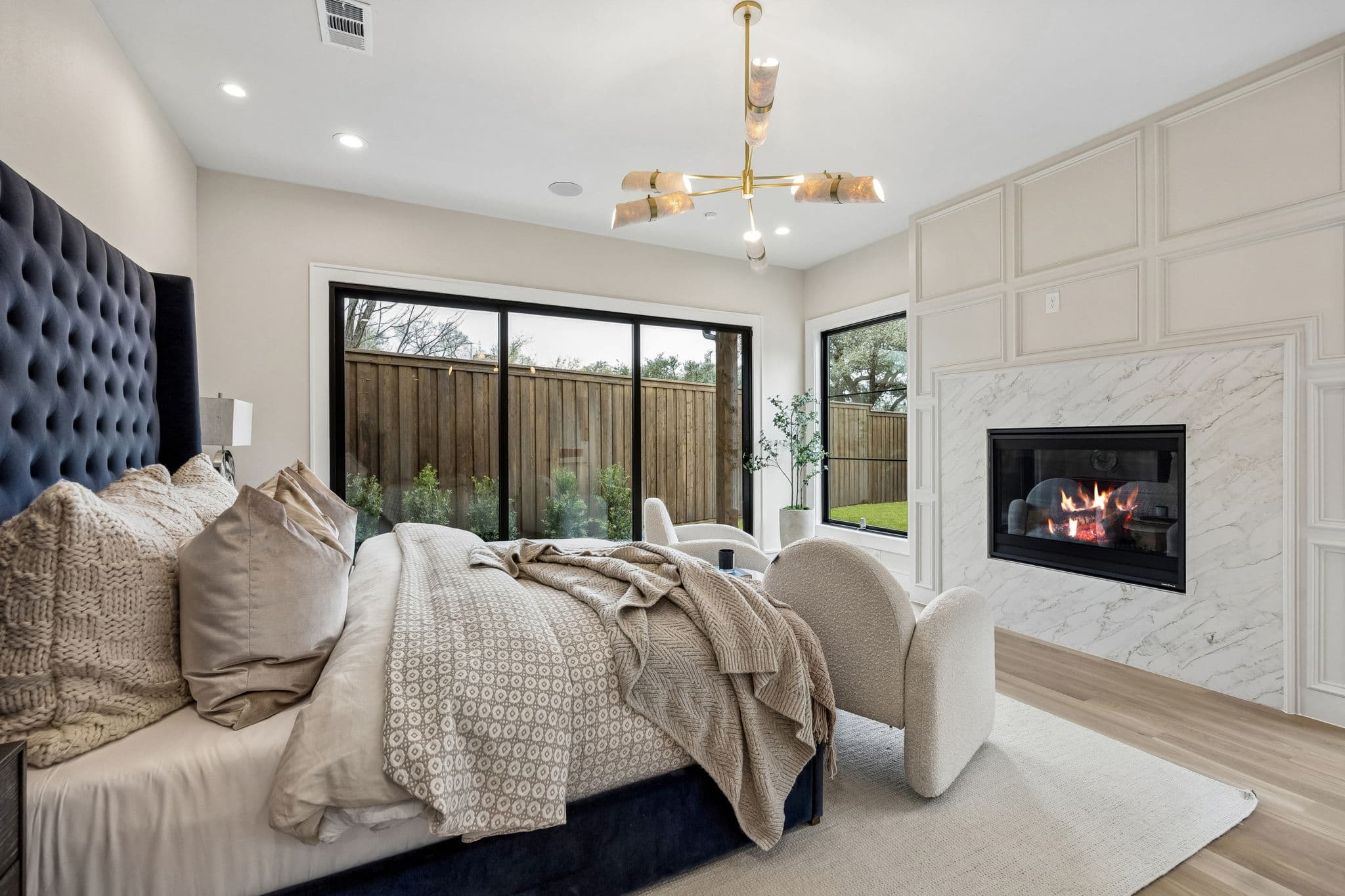 The primary bedroom at 7272 Tangleglen, a navy buttoned headboard the width of the wall, a fireplace beside it and doors onto the garden.