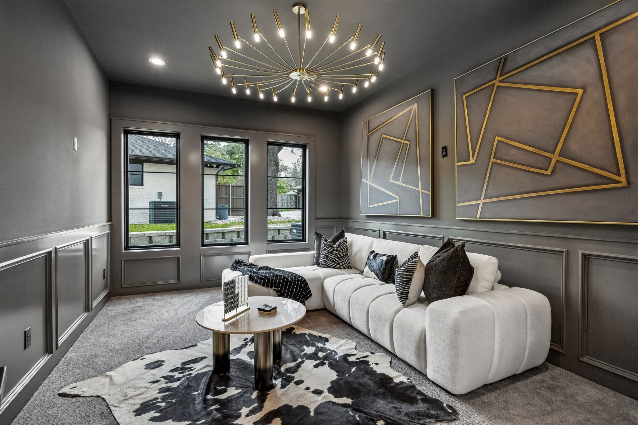The sitting room at 7272 Tangleglen, charcoal panelling inlaid with brass, a pale sectional on a hide rug and a burst chandelier overhead.